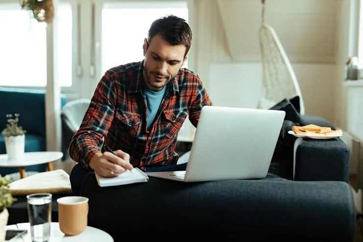 A man writing notes while working on a computer at home