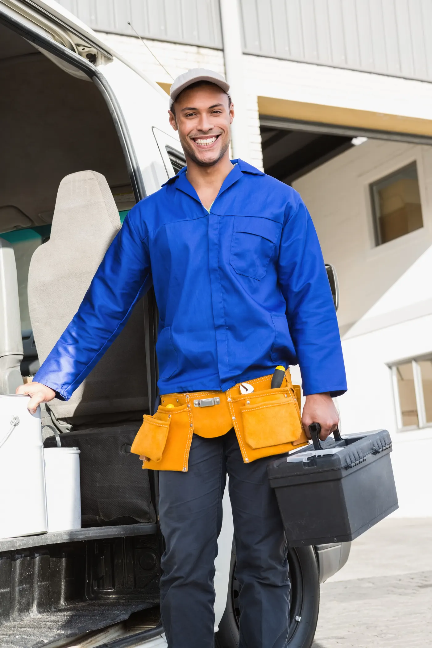 A smiling handyman with a tool belt and toolbox standing beside his van